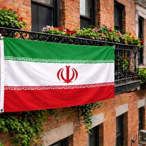 Puede incluir: Una bandera iraní, con franjas horizontales verdes, blancas y rojas, cuelga de un balcón de hierro forjado negro. La bandera presenta un emblema rojo central y escritura árabe en los bordes. El balcón está adornado con flores y vegetación, sobre un edificio de ladrillo.