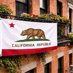 Puede incluir: Una bandera de la República de California con un oso pardo, una estrella roja y una franja roja. La bandera se muestra en un balcón de hierro forjado con flores y vegetación, contra un edificio de ladrillo.