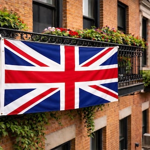 May include: A rectangular Union Jack flag featuring a blue background with a white-bordered red cross and diagonal red stripes. The flag is displayed horizontally, attached to a black metal balcony railing on a brick building exterior.