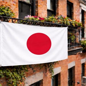 Könnte beinhalten: Eine weiße Flagge mit einem großen roten Kreis in der Mitte, die Flagge Japans, hängt an einem Balkon. Die Flagge wird vor dem Hintergrund eines Backsteingebäudes mit Fenstern und Blumenarrangements gezeigt.