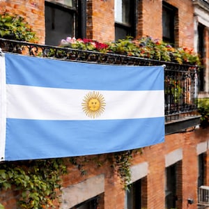Pode incluir: A bandeira nacional da Argentina composta por três faixas horizontais em azul-claro e branco com um emblema solar dourado, o Sol de Maio, centralizado na faixa branca. A bandeira é exibida horizontalmente contra uma fachada de tijolos.