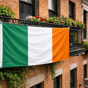 Puede incluir: Una bandera rectangular con tres franjas verticales de igual anchura en verde, blanco y naranja. La bandera se muestra horizontalmente, sujeta a una barandilla de balcón de metal negro con ojales visibles en las esquinas y una textura de tela lisa.