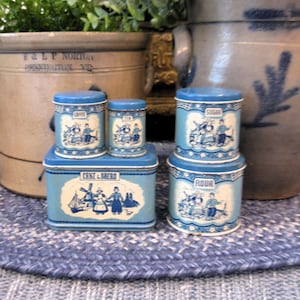 May include: Five blue and white ceramic canisters with lids. The canisters are decorated with a Dutch theme and have the words "Cake & Sugar", "Flour", "Tea", and "Sugar" printed on them.