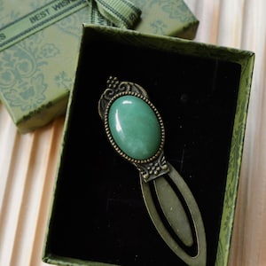 May include: A green gemstone bookmark with an oval stone set in an ornate, antique-style bronze frame. The bookmark is presented in a green gift box with a matching ribbon that reads "BEST WISHES".