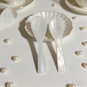 May include: Two pearlescent white mother-of-pearl spoons with a subtle iridescent sheen, resting on a white scalloped plate with a gold rim. Scattered pearls surround the spoons and plate, suggesting a theme of elegance and refinement.