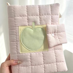 May include: A light pink quilted notebook cover featuring a square patch with a pale green apple appliqu&eacute;. The cover includes a small attached pocket on the right side with a white star-shaped stud and a white star-shaped zipper pull at the top.