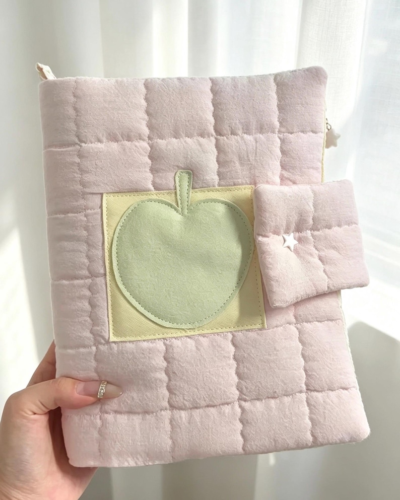 May include: A light pink quilted notebook cover featuring a square patch with a pale green apple appliqu&eacute;. The cover includes a small attached pocket on the right side with a white star-shaped stud and a white star-shaped zipper pull at the top.