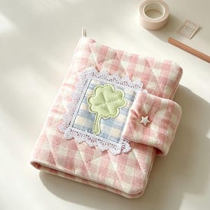 May include: A quilted fabric notebook cover featuring a pink and white gingham pattern. The front displays a square patch with a light green four-leaf clover appliqué, framed by white lace trim. A matching tab closure includes a small white star button.