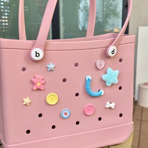 May include: A pale pink tote bag with matching handles and decorative charms. The bag features various charms, including stars, hearts, buttons, and a teddy bear. The bag has the letter "b" on the handle attachments.