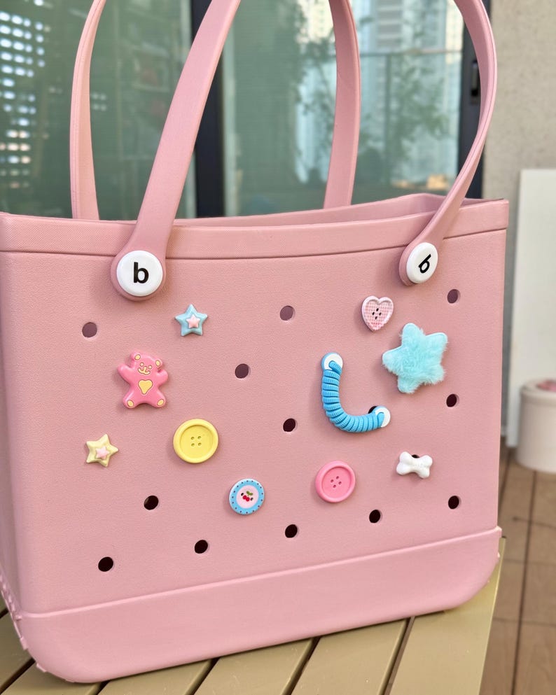 May include: A pale pink tote bag with matching handles and decorative charms. The bag features various charms, including stars, hearts, buttons, and a teddy bear. The bag has the letter "b" on the handle attachments.