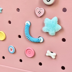 May include: A selection of decorative charms and buttons in various shapes and colours, including a fluffy blue star, a pink heart-shaped button, and a blue coiled charm. The items are arranged on a pink surface with evenly spaced holes.