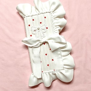 May include: A white, rectangular case with a ruffled border and a large bow. The case features a decorative bow and small red hearts. The design is symmetrical, with a pearl accent on the bow. The background is a solid pink.