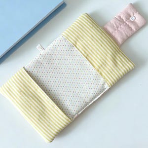 May include: A rectangular fabric pouch featuring a white centre panel with a multicoloured polka dot pattern, flanked by yellow and white striped fabric sections. A pale pink fabric flap with a white snap closure secures the top opening.