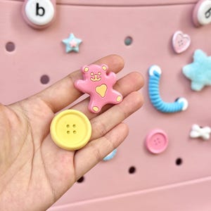 May include: A selection of colourful decorative charms and buttons. Includes a yellow button with four holes, a pink teddy bear, a blue swirly shape, and a pink heart. The background is a light pink colour.