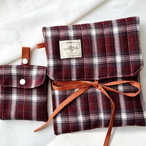 May include: Two-piece set featuring a burgundy, white, and gray plaid pattern. The set includes a small pouch with a button closure and a larger bag with a flap and a brown ribbon tie. A small label is sewn on the larger bag.