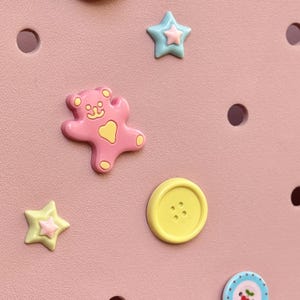 May include: A close-up of a pink surface adorned with decorative embellishments. Features a pink bear, yellow button, and star-shaped charms in yellow and blue. The background has evenly spaced circular holes.