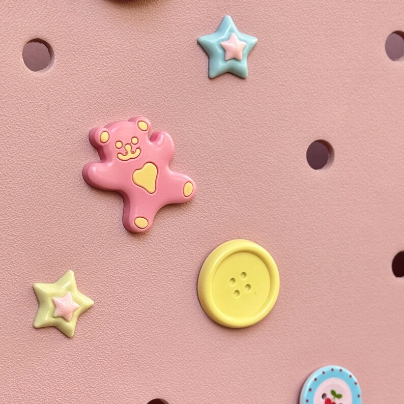 May include: A close-up of a pink surface adorned with decorative embellishments. Features a pink bear, yellow button, and star-shaped charms in yellow and blue. The background has evenly spaced circular holes.