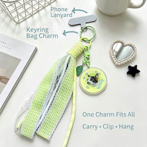 Puede incluir: Un accesorio de borla de tela de cuadros vichy verde lima y blanco con un dije circular que muestra una ilustración de un gato y el texto "Spring". El accesorio incluye un llavero verde claro, un dije de hoja pequeña y un cordón amarillo.