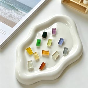 May include: A white, organically shaped ceramic tray holds an assortment of small, rectangular glass tiles in various colors. The tiles include shades of yellow, green, orange, purple, and gray. The tray sits on a white surface, with a magazine and small wooden shelf in the background.