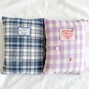 May include: Two decorative cushions with a plaid pattern. One is blue and white, with a "Lucky Cat" patch. The other is lavender and white, with a "Find your love" patch and buttons.