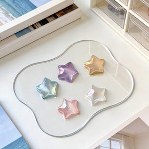 May include: A clear, wavy-edged tray holds five iridescent star-shaped decorations in shades of blue, purple, pink, gold, and white. The tray sits on a white surface, with books and a small storage unit in the background.