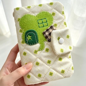May include: A quilted fabric pouch in cream with a repeating green clover pattern. The front features a large green felt clover appliqué shaped like a house with a window and a dark green door with a star. A small flap with a white snap closure.