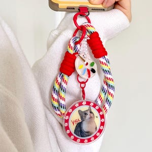 Peut inclure: Une dragonne de téléphone en cordon blanc à motif arc-en-ciel, avec des accents en cordon rouge et un clip en plastique rouge. Deux breloques sont attachées : une palette de peinture blanche avec des taches colorées et un disque avec un chat et le texte YSL.