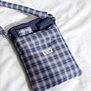 May include: A blue plaid crossbody bag with a matching strap. The bag features a navy blue interior and a small rectangular pocket. A small label with text and a graphic is sewn on the front.