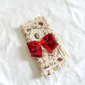 May include: A rectangular fabric case featuring a cream background with a red floral pattern, edged with cream-colored lace trim. The center is adorned with a white lace doily, a small oval cameo brooch, and a prominent dark red satin ribbon bow.
