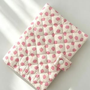 May include: A quilted fabric cover featuring a repeating pattern of small pink strawberries and tiny brown dots on a cream background. The cover has a diamond-stitched texture and includes a small tab closure secured with a single white snap button.