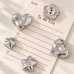 May include: A cream-coloured paper notepad with the text "DATE" and "To do list" printed in red, featuring five silver-toned metallic magnets in the shapes of two hearts, two stars, and one flower with two red dots in the centre.