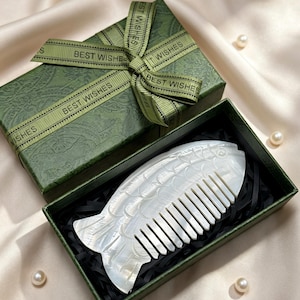 May include: A fish-shaped comb made of mother-of-pearl, presented in a green gift box with a matching ribbon that reads "BEST WISHES". The comb is white and has detailed scales. The box is on a satin surface.