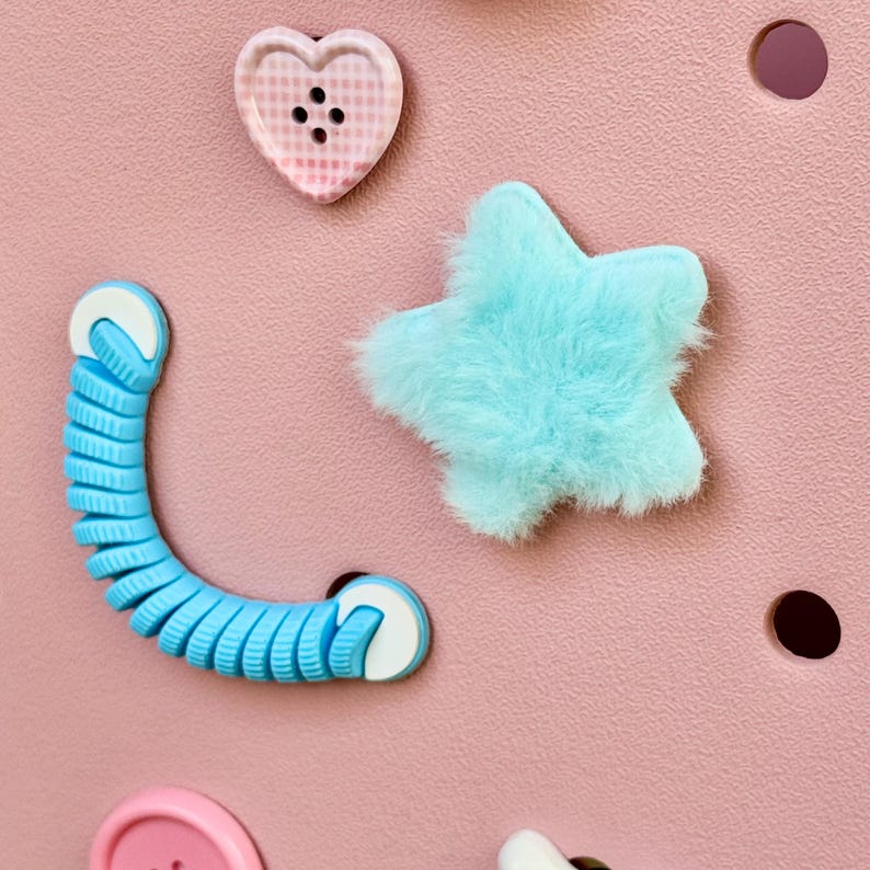 May include: A close-up of a pink surface with various decorative elements. A light blue, fluffy star and a blue, coiled, flexible cord are visible. A pink heart-shaped button and other buttons are also present.