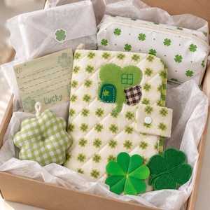 May include: A gift box containing a quilted notebook with a clover house appliqué, a white zippered pouch with green clover patterns, a green and white gingham clover-shaped pin cushion, a clover-themed greeting card with the word Lucky, and patches.
