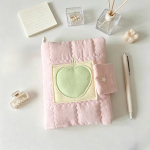 May include: A light pink quilted notebook cover featuring a central square patch with a light green apple appliqu&eacute;. The cover includes a small side flap secured with a white star-shaped button, providing a soft, textured appearance for a standard journal.
