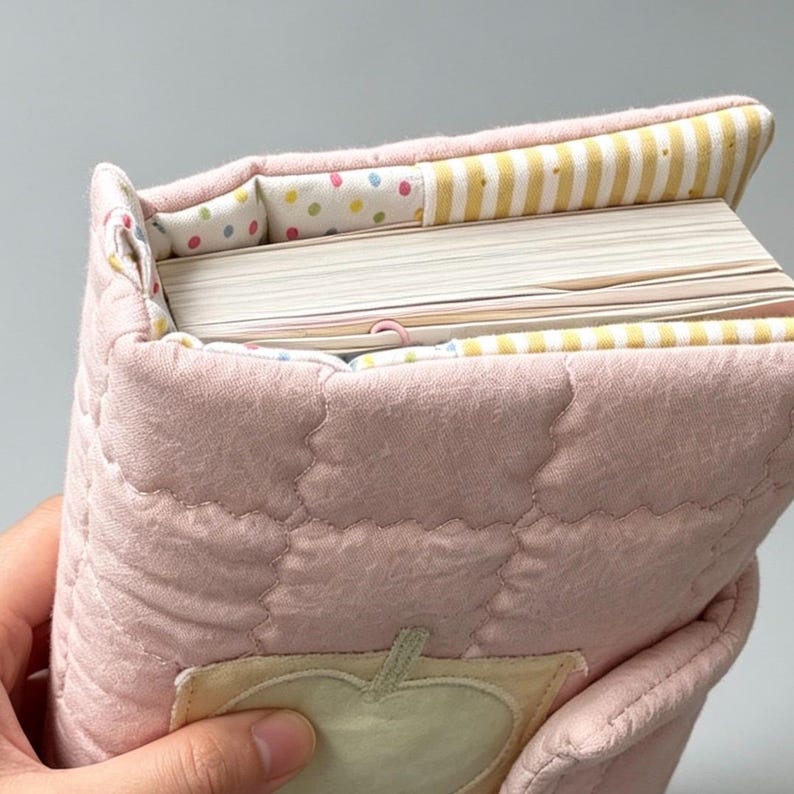 May include: A quilted pink fabric book cover featuring a light yellow apple appliqu&eacute; on the front. The interior lining displays a pattern of colourful polka dots and yellow and white stripes, enclosing a thick book with white pages.