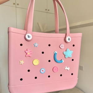 May include: A pink tote bag with two handles and decorative charms. The bag has a series of holes and is adorned with various charms, including stars, buttons, and a teddy bear. The bag has the letter "b" on both sides.