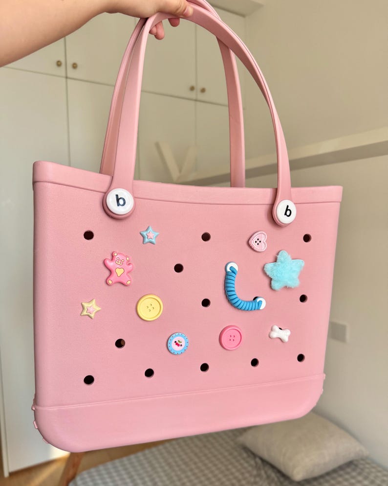 May include: A pink tote bag with two handles and decorative charms. The bag has a series of holes and is adorned with various charms, including stars, buttons, and a teddy bear. The bag has the letter "b" on both sides.