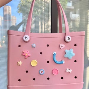 May include: A pale pink tote bag with two handles and decorative charms. The bag has a series of holes and is decorated with star, heart, and button-shaped embellishments. The handles are attached with white circular fasteners with a small "b" on them.