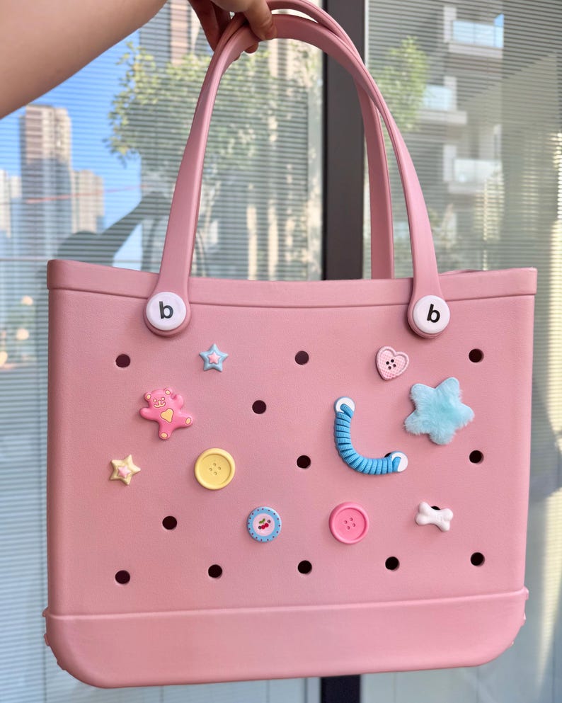 May include: A pale pink tote bag with two handles and decorative charms. The bag has a series of holes and is decorated with star, heart, and button-shaped embellishments. The handles are attached with white circular fasteners with a small "b" on them.