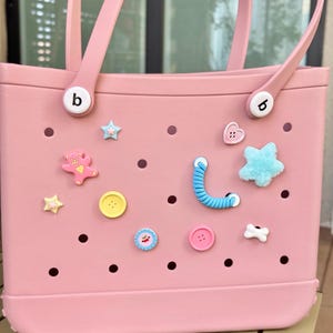 May include: A light pink tote bag with two handles and decorative charms. The bag has a rectangular shape with a flat bottom and is adorned with various charms, including stars, hearts, and buttons. The bag has the letter "b" on the side.