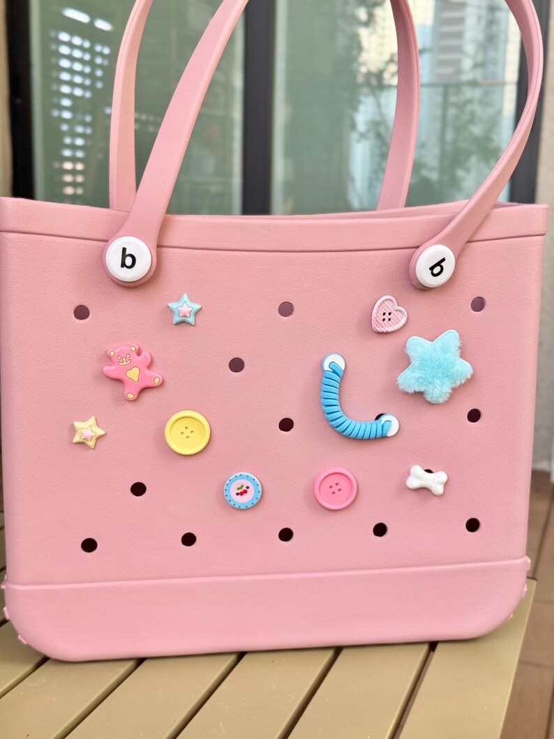 May include: A light pink tote bag with two handles and decorative charms. The bag has a rectangular shape with a flat bottom and is adorned with various charms, including stars, hearts, and buttons. The bag has the letter "b" on the side.