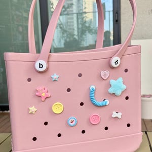 May include: A pale pink tote bag with matching handles and decorative charms. The bag features various charms, including stars, hearts, buttons, and a teddy bear. The bag has a rectangular shape and is made of a durable material.