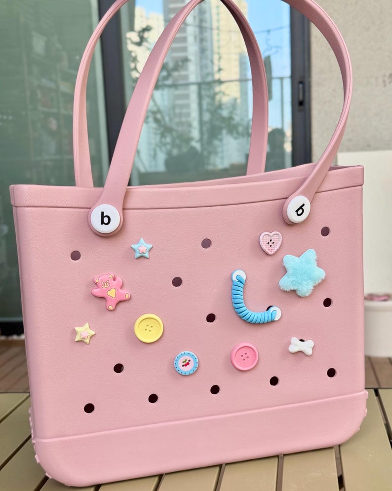 May include: A pale pink tote bag with matching handles and decorative charms. The bag features various charms, including stars, hearts, buttons, and a teddy bear. The bag has a rectangular shape and is made of a durable material.
