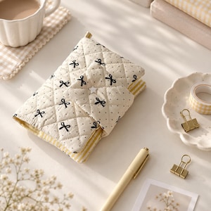 May include: A quilted fabric notebook cover in cream featuring a black polka dot and bow pattern. The cover has a diamond-stitched texture, a fold-over flap secured with a small white star-shaped button, and reveals yellow and white striped lining.
