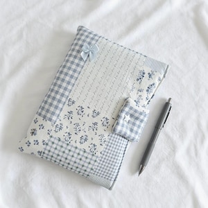 Könnte beinhalten: Ein Patchwork-Notizbuchumschlag in Blau- und Weißtönen mit Blumen- und Vichy-Mustern. Eine kleine blaue Schleife ziert die Oberseite. Ein passender Stift liegt neben dem Notizbuch, das eine kleine Tasche mit einem Stern hat. Das Notizbuch liegt auf weißem Hintergrund.