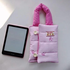 May include: A pink and white striped Kindle sleeve with a matching handle and button closures. The sleeve has a small patch with three bear illustrations and the text "Sweet Bear". A Kindle e-reader is next to the sleeve.