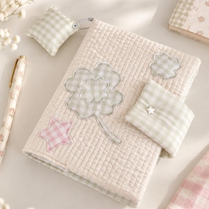 May include: A quilted fabric notebook cover in cream featuring a large sage green gingham four-leaf clover appliqué, a small pink gingham star, and a cloud shape. It includes a matching sage green gingham strap with a small silver star stud.