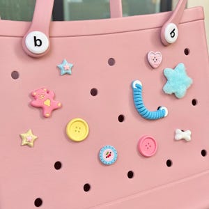 May include: Pink tote bag with various decorative charms. Charms include a pink teddy bear, stars, buttons, a blue worm, a heart, and a bone. The bag has two handles and the letter "b" on one side.