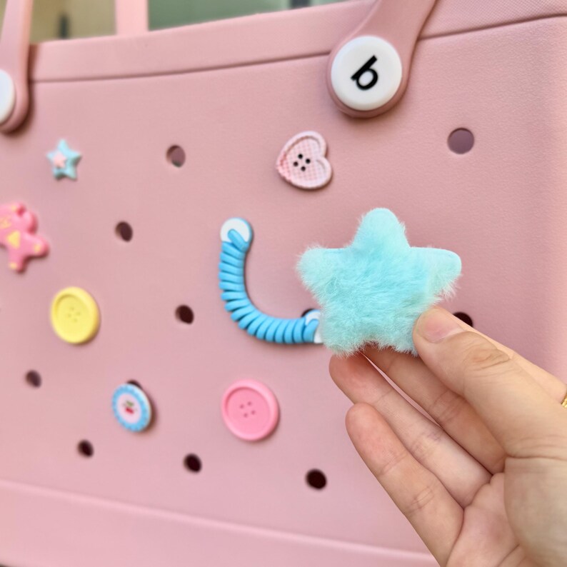 May include: A pale pink tote bag adorned with decorative elements. The bag includes a light blue fluffy star, a blue coiled detail, and various button-shaped charms in different colours. The bag has a handle with a white button.